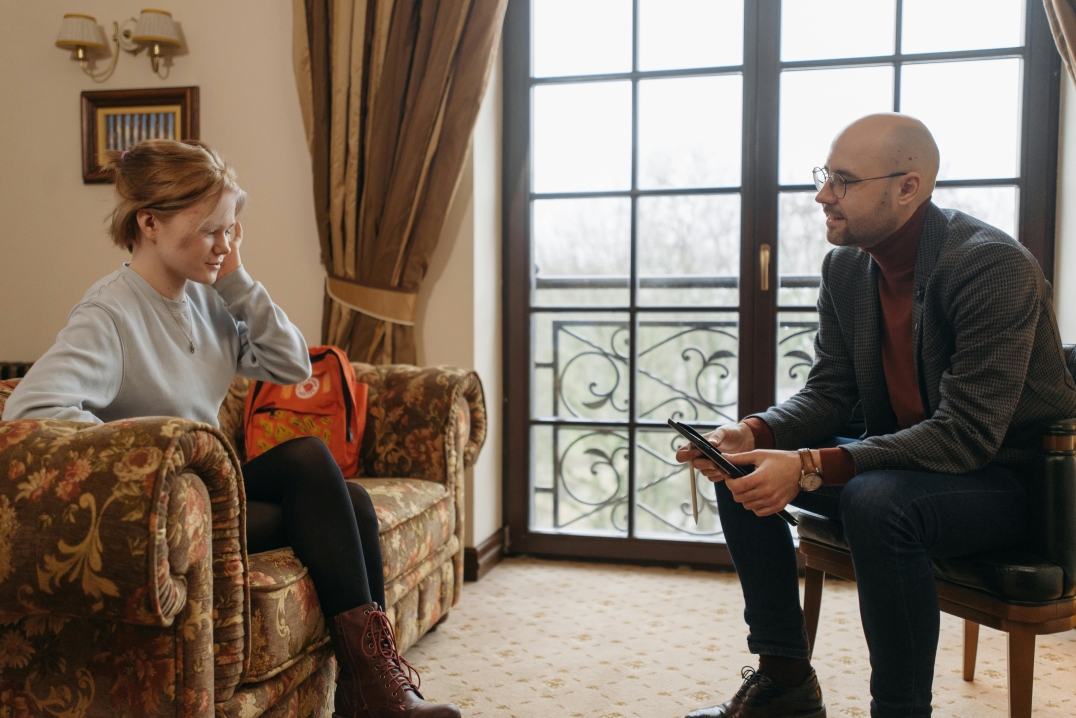 Jongere en begeleider zitten tegenover elkaar in gesprek in een rustige, huiselijke omgeving.